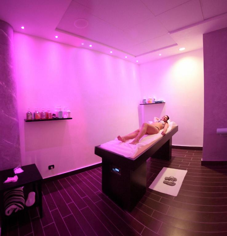 Relaksējoša spa telpa ar rozā apgaismojumu, kurā atpūšas sieviete uz masāžas galda. Uz plaukta redzami dažādi kosmētikas līdzekļi un dvieļi.