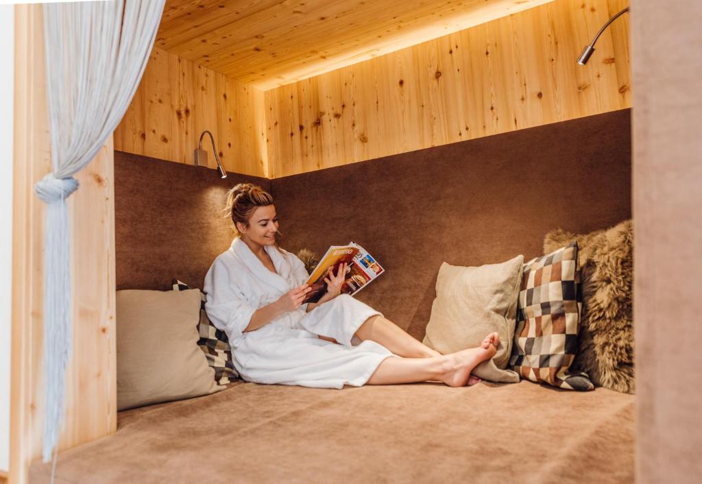 Sieviete baltā halātā atpūšas mājīgā koka telpā, lasot grāmatu un sēžot uz mīkstas dīvāna. Telpa ir apgaismota ar siltu gaismu, radot relaksējošu atmosfēru.