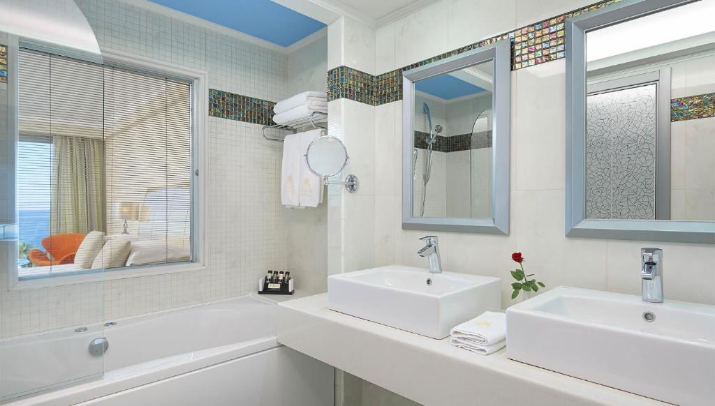 Mūsdienīga vannas istaba ar diviem izlietnēm, lieliem spoguļiem un logu, caur kuru redzama viesnīcas istaba. Interjers ir gaišs ar krāsainām flīzēm un dekoratīvu rožu uz galda.