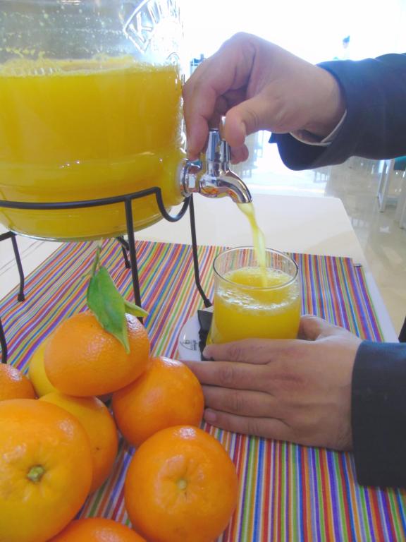 Svaiga apelsīnu sula tiek ielejama no trauka glāzē, blakus redzami apelsīni uz krāsainas galdautas.