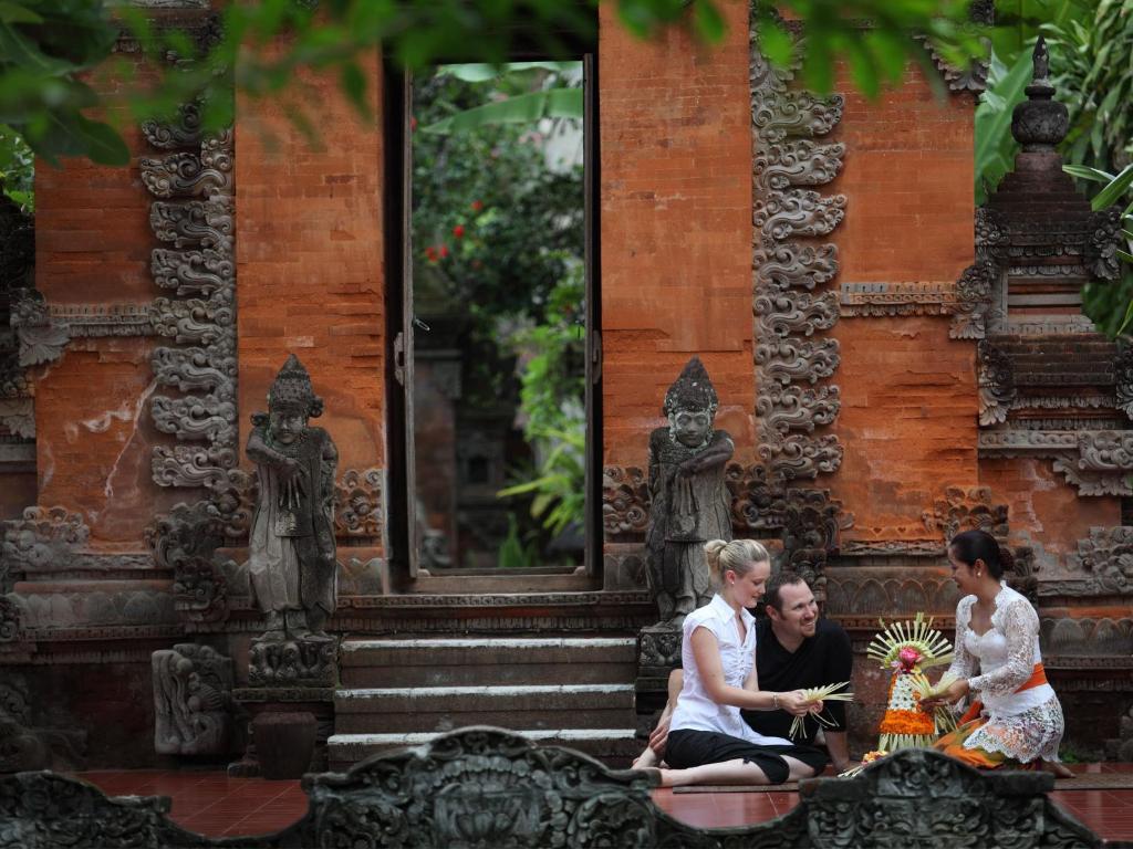 Trīs cilvēki, tostarp divi tūristi un vietējā sieviete, piedalās tradicionālajā ceremonijā, veidojot ziedu un lapu kompozīcijas pie baliešu tempļa ar izsmalcinātām akmens skulptūrām un sarkanu sienu fona.
