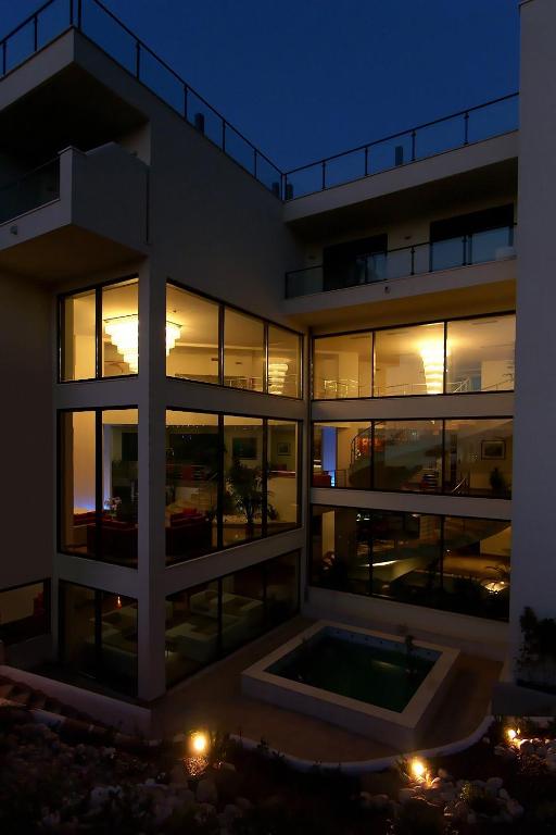 Mūsdienīga māja ar plašiem logiem, kas izgaismota naktī, radot elegantu atmosfēru. Redzams baseins un terase, kas papildina mājas dizainu.