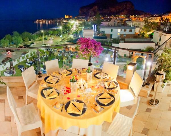 Elegantā vakariņu galda uzstādījums ar ziediem, kas atrodas terasē pie jūras, ar skatu uz apgaismoto pilsētu naktī. Galda klājums ietver melnas šķīvjus un dekoratīvus elementus, radot romantisku atmosfēru.