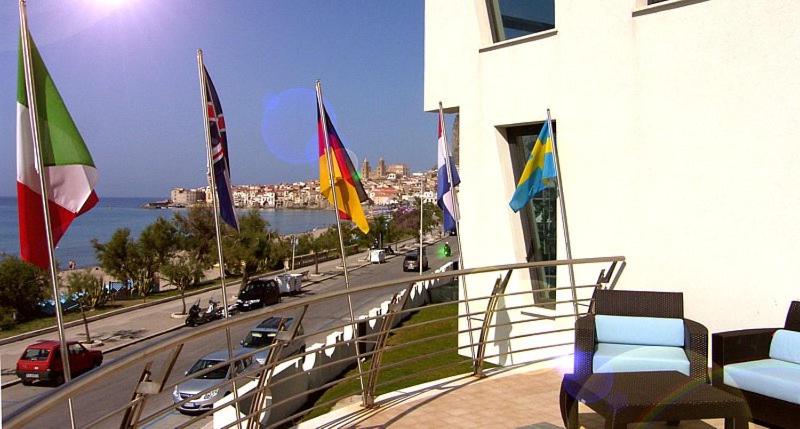 Balkons ar skatu uz jūru un plīvojošām karogiem, tostarp Itālijas, Vācijas un Lielbritānijas, izceļot starptautisku atmosfēru. Diena ir saulaina, radot patīkamu noskaņu.