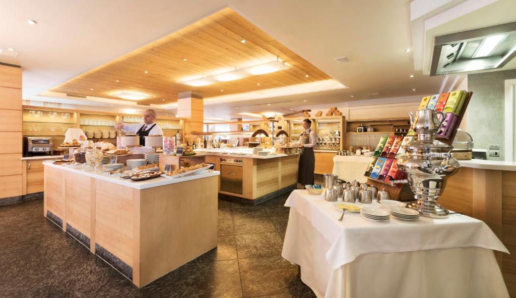 Elegants restorāna interjers ar bufetes galdu, kas pilns ar dažādiem ēdieniem un dzērieniem, un apkalpojošais personāls. Gaisma un koka elementi rada siltu atmosfēru.