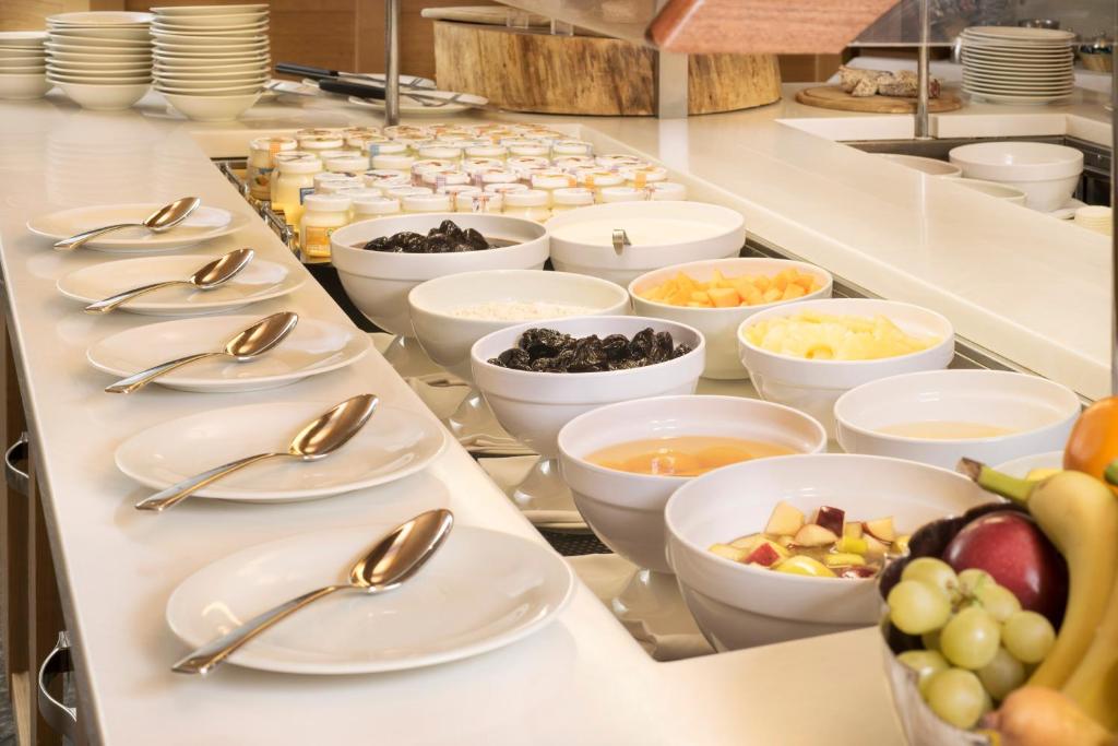 Brokastu bufete ar dažādiem augļiem, jogurtiem un graudaugiem, kas pasniegti baltos traukos un ar karotēm. Galda dizains ir moderns un sakārtots, radot patīkamu atmosfēru.