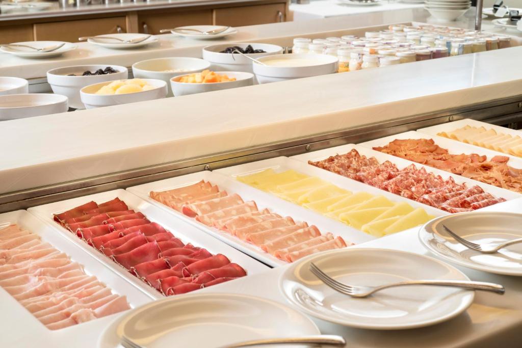 Svaigu gaļas un sieru izkārtojums uz bufetes galda, piedāvājot plašu izvēli. Fonā redzami dažādi augļi un jogurtu trauki, radot gardu brokastu atmosfēru.