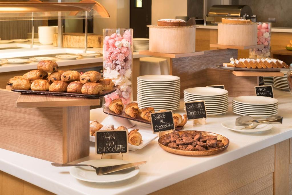 Bufete ar dažādiem konditorejas izstrādājumiem, tostarp šokolādes kruasāniem, cepumiem un kūkām, izkārtota uz elegantām koka virsmām. Galdiņā redzami arī balti šķīvji un saldumi.