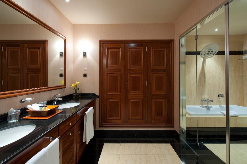 Moderns vannas istabas interjers ar melnu akmens izstrādājumu, koka skapjiem un divām izlietnēm. Gaismas elementi rada siltu atmosfēru, un stikla duša piešķir elegantumu.