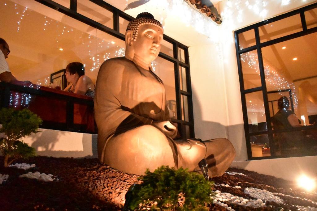 Buda statuja atrodas apgaismotā telpā, apņemta ar zaļiem augiem un dekoratīviem akmeņiem. Fonā redzami cilvēki, kas bauda ēdienu un sarunas.