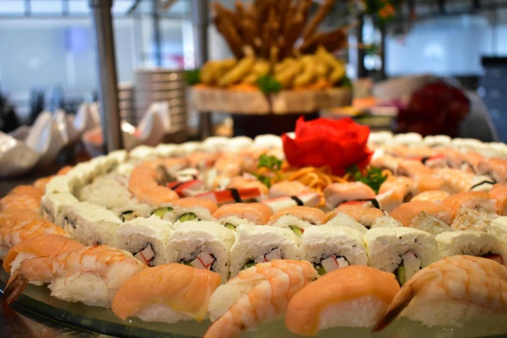 Svaigu sushi un garneles, sakārtotas apļveida kompozīcijā, rotātas ar sarkano ziedu un zaļumiem. Ideāla izvēle sushi mīļotājiem.