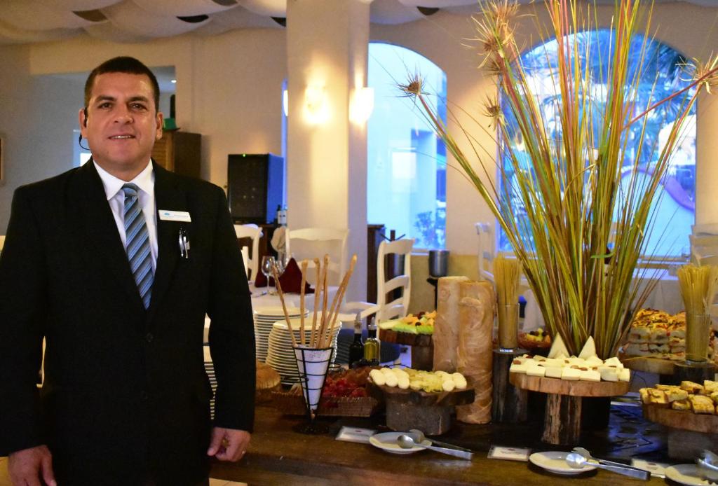 Sakopta restorāna darbinieks stāv blakus bagātīgai ēdienu izvēlei, kas izkārtota uz koka galda. Fons ir gaiši apgaismots ar logiem, kas sniedz skatu uz ārpusi.