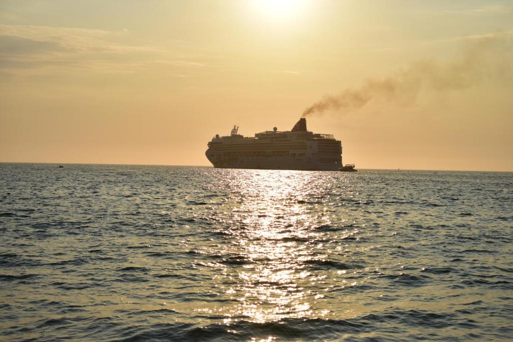 Kruīza kuģis ceļo pa saulrieta apmirdzētu jūru, atstājot dūmu pēdas gaisā. Ūdens virsma atspoguļo saules gaismu, radot gleznainu skatu.