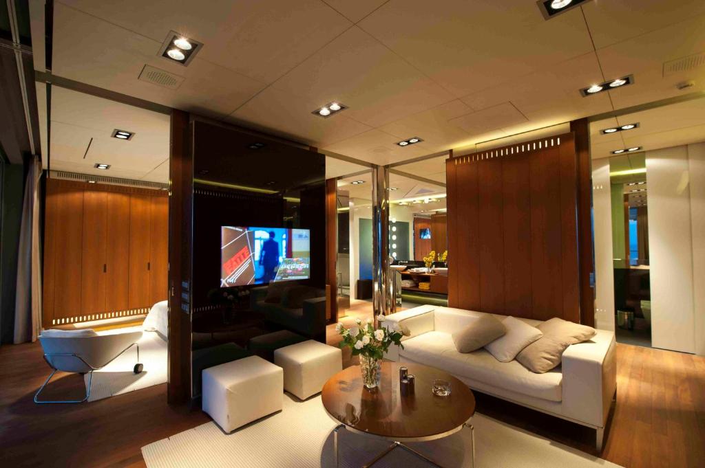 Mūsdienīgs un elegants viesistabas interjers ar gaišiem dīvāniem, koka paneļiem un mūsdienīgu televizoru. Telpā ir daudz apgaismojuma un dekoratīvu elementu, kas rada mājīgu atmosfēru.