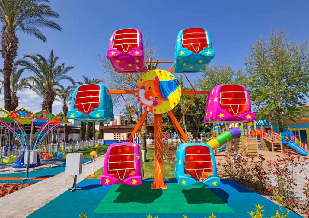 Krāsains ritenis ar kabīnēm bērnu atrakciju parkā, ko ieskauj palmām un ziediem. Šis jautrais ritenis ir lieliska izklaide ģimenēm ar bērniem.