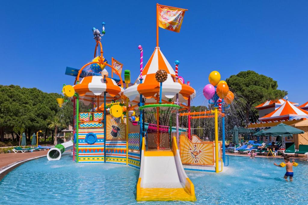 Krāsains ūdens atrakciju parks ar slidkalniņu un strūklakām, bērni izklaidējas ūdenī saulainā dienā. Fons ietver kokus un sauļošanās krēslus.