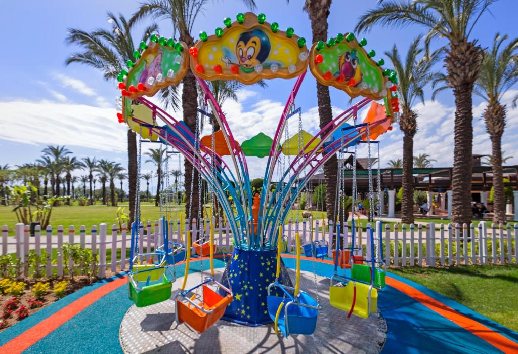 Krāsains bērnu karuselis ar šūpuļiem, izvietots saulainā, zaļā parkā ar palmām un ziediem. Ideāla vieta bērnu izklaidei un priecīgām atmiņām.