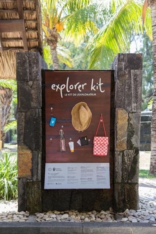 Reklāmas stends ar tekstu "explorer kit", kas ietver dažādus ceļojumu piederumus, piemēram, cepuri, ūdens pudeli un somu, fona palmām un dārza ainavu.