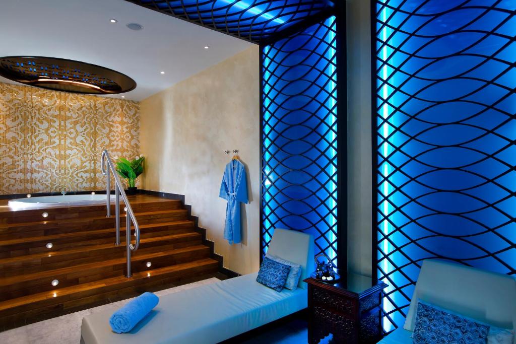 Mūsdienīgs spa interjers ar koka kāpnēm, zilām apgaismojuma detaļām un relaksējošām atpūtas zonām. Sienas rotā eleganta mozaīkas apdare un augi, radot mierīgu atmosfēru.
