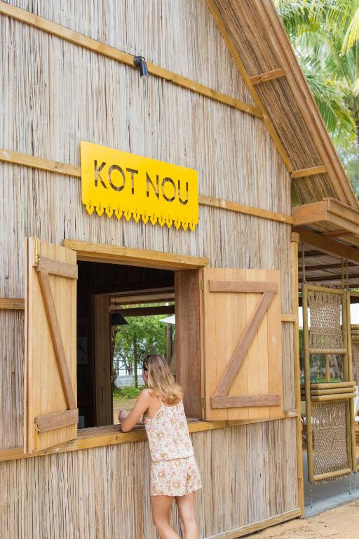 Bārda ar dzeltenu zīmi "KOT NOU", kura ir izgatavota no dabīgiem materiāliem un atrodas pie ūdens. Sieviete apmeklē bāru, izskatoties pēc atpūtas brīža.