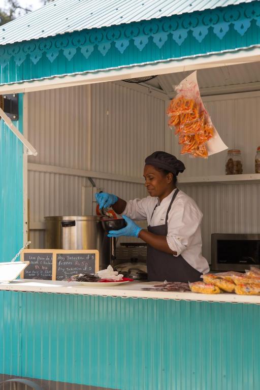 Sieviete, kas gatavo ēdienu, strādā gaišā, zilā ēdienu stendā. Uz galda ir šķīvji ar pārtiku un cena, kā arī karstā katlā vārās ēdiens.