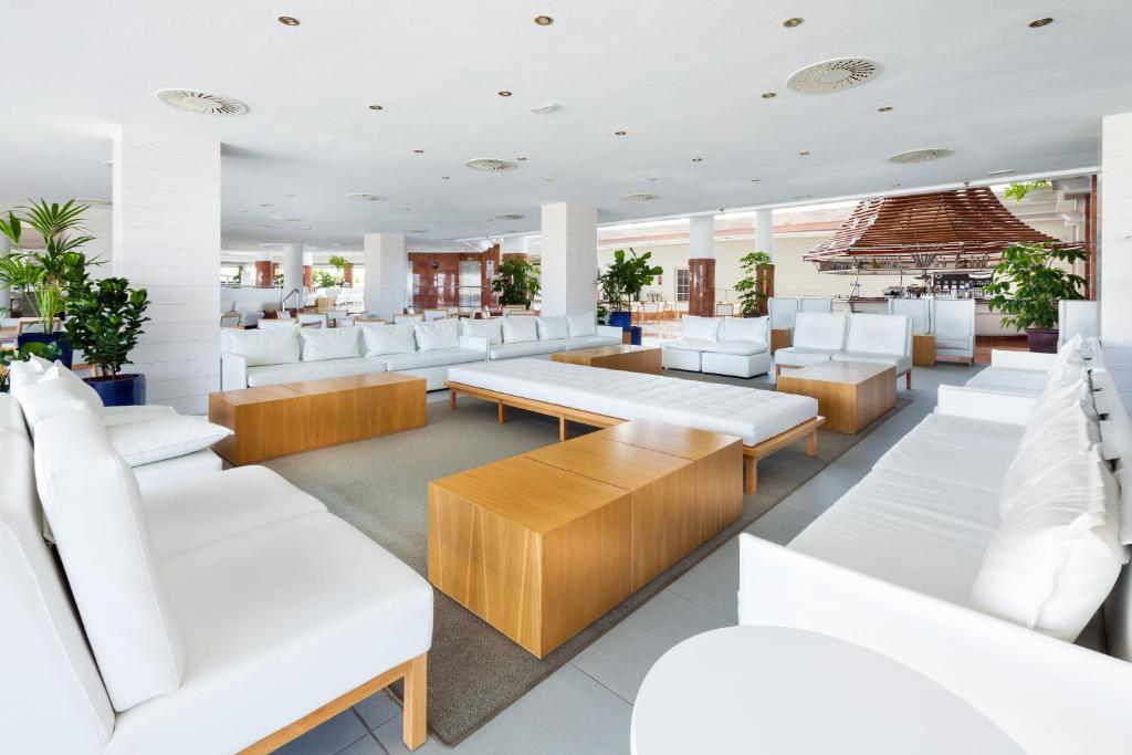 Mūsdienīgs viesnīcas atpūtas telpa ar baltām mēbelēm un koka galdiem, kas ieskauta zaļiem augiem. Telpa ir gaiša un plaša, ideāli piemērota relaksācijai.