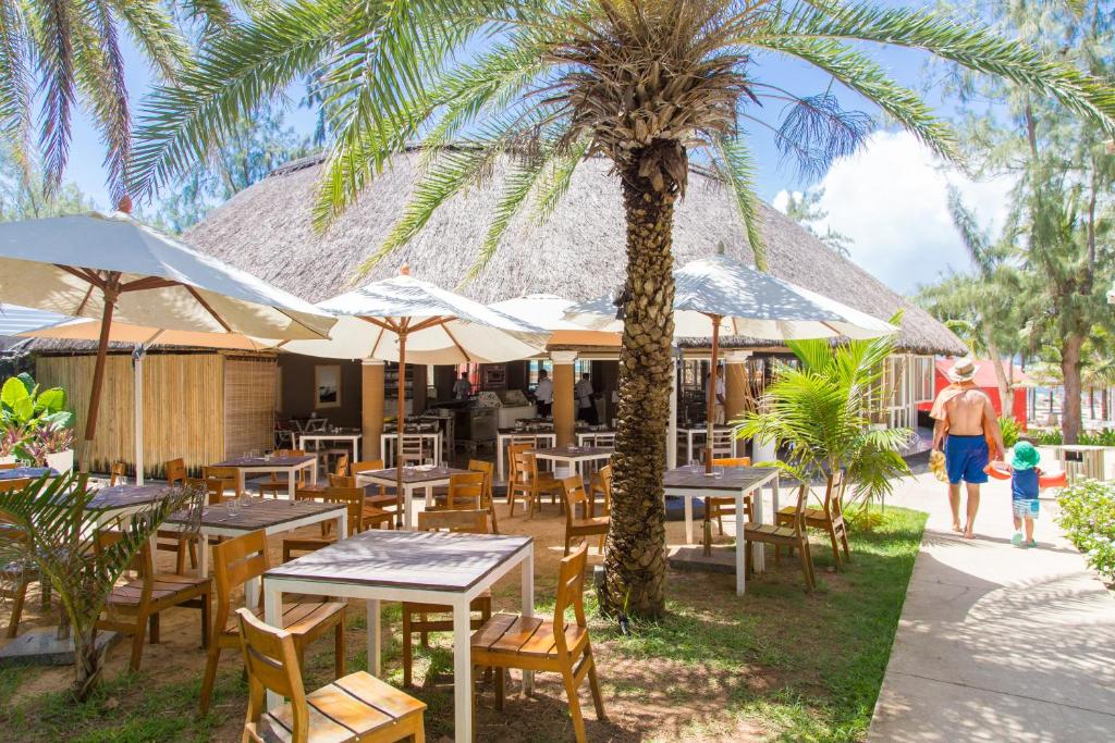 Tropu restorāns ar brīvdabas terasi, kurā ir koka galdi un krēsli, palmām un saules parasoliem. Aiz skatu punkta redzams cilvēks ar bērnu, dodoties uz pludmali.