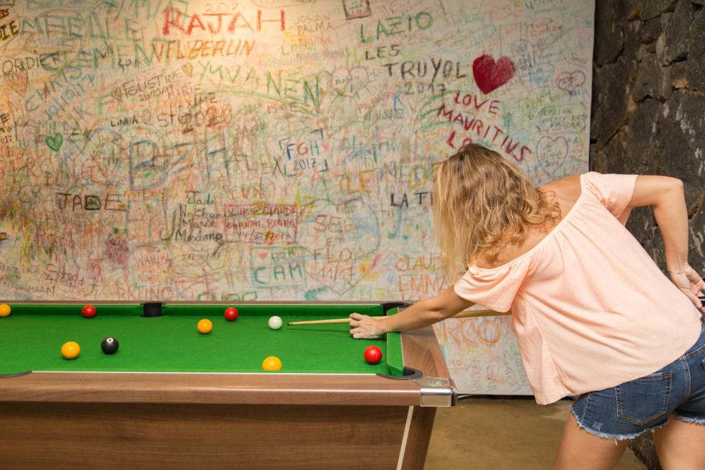 Sieviete spēlē biljardu pie galda ar zaļu virsmu, fonā redzama krāsaina siena, kas klāta ar dažādiem grafiti.
