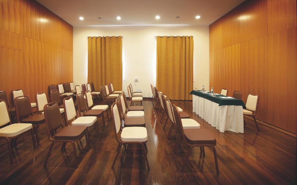 Konferenču telpa ar sēdekļiem, kas sakārtoti rindās, un galdu priekšā ar dzeramā ūdens pudelēm. Telpas interjers ir moderns ar koka apdari un aizkariem.