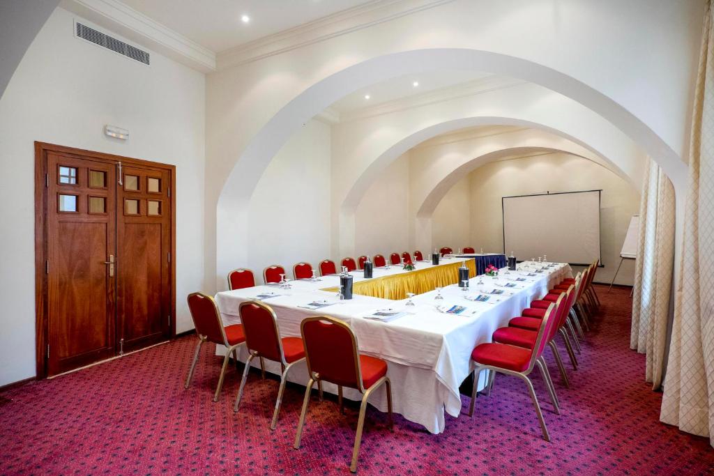 Konferenču telpa ar garu galdu un sarkaniem krēsliem, kurā redzamas prezentācijas iekārtas un organizēti galda piederumi. Telpa ir gaiša, ar arku formas griestiem un siltu interjeru.