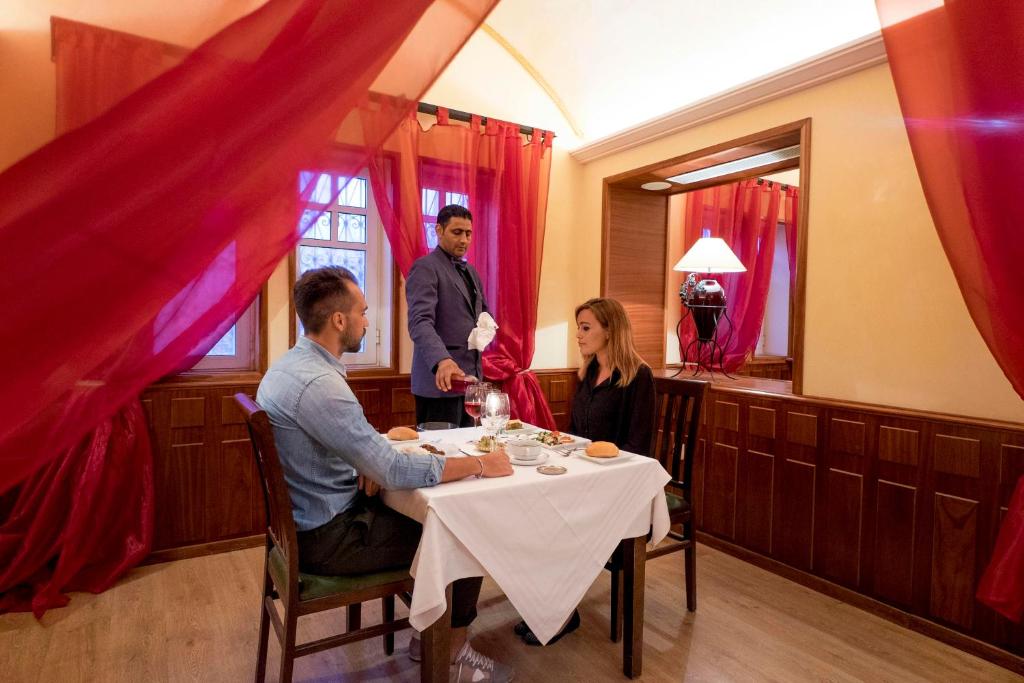 Romantiska vakariņas restorānā ar sarkaniem aizkariem, kur viesmīlis apkalpo pāri pie galda.