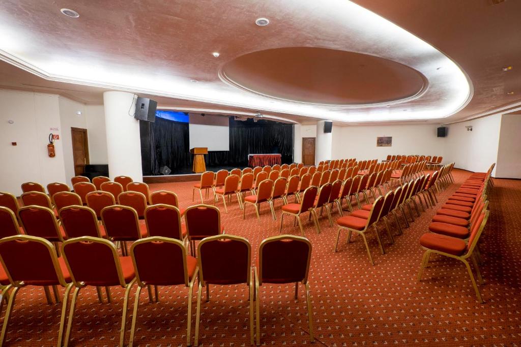Konferenču zāle ar sarkaniem krēsliem, sakārtotiem rindās, gatava pasākumiem. Aizmugurē redzams melns aizkars un skatuve ar lektora galdu.