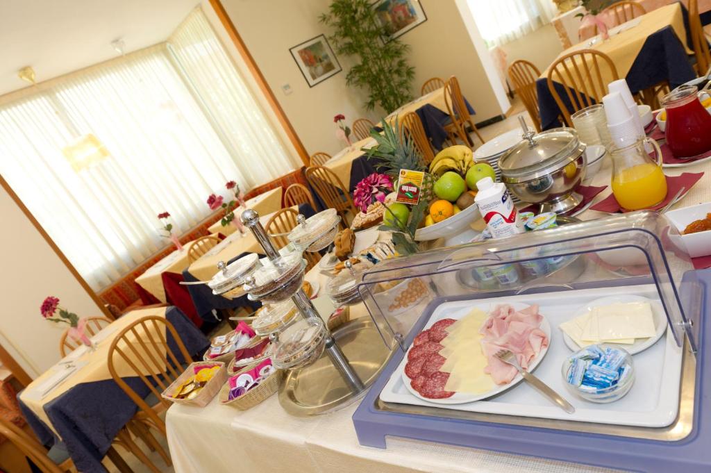 Brokastu galds ar dažādām delikatesēm, augļiem un dzērieniem, kas piedāvā daudzveidīgu izvēli. Gaisīga atmosfēra ar koka mēbelēm un ziediem uz galdiem.