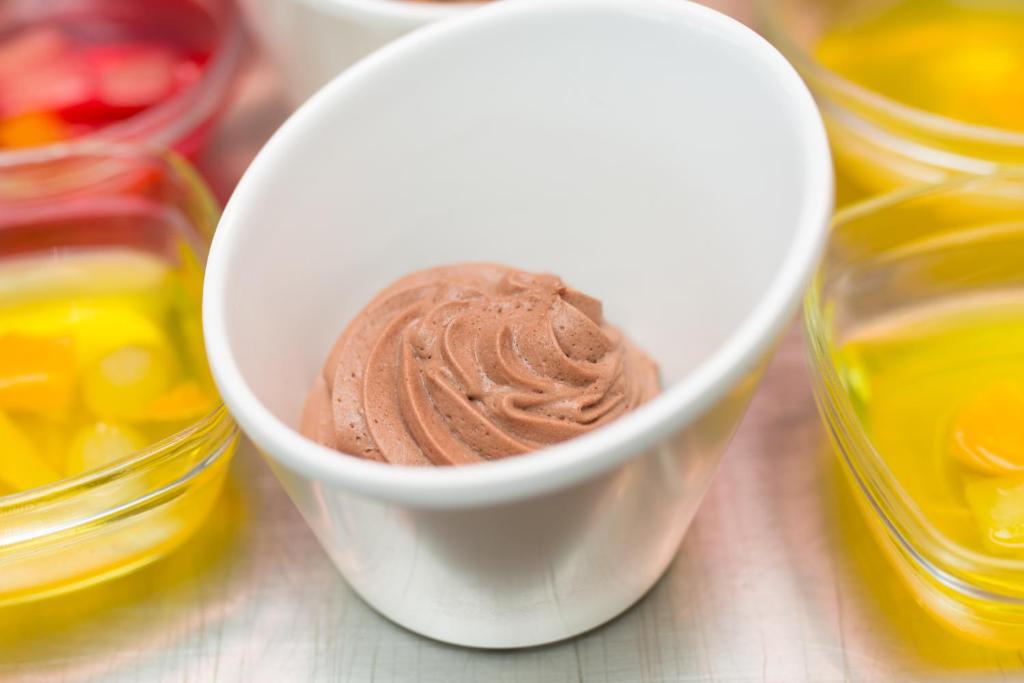 Šajā attēlā redzama šokolādes krēma porcija baltā traukā, ko ieskauj dažādu krāsu želejas un augļu gabaliņi. Krēms ir gludi sakults, radot pievilcīgu tekstūru.