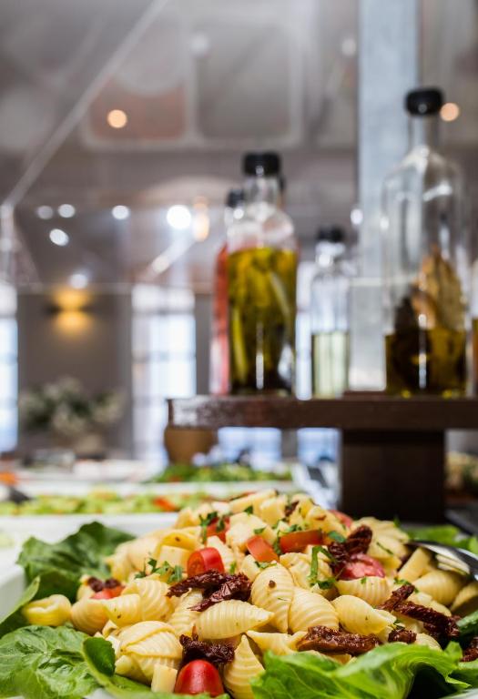 Pasta salāti ar saulē žāvētiem tomātiem un svaigiem dārzeņiem, pasniegti uz salātu lapām. Fons ar glāzēm un zālēm, radot elegantu atmosfēru.
