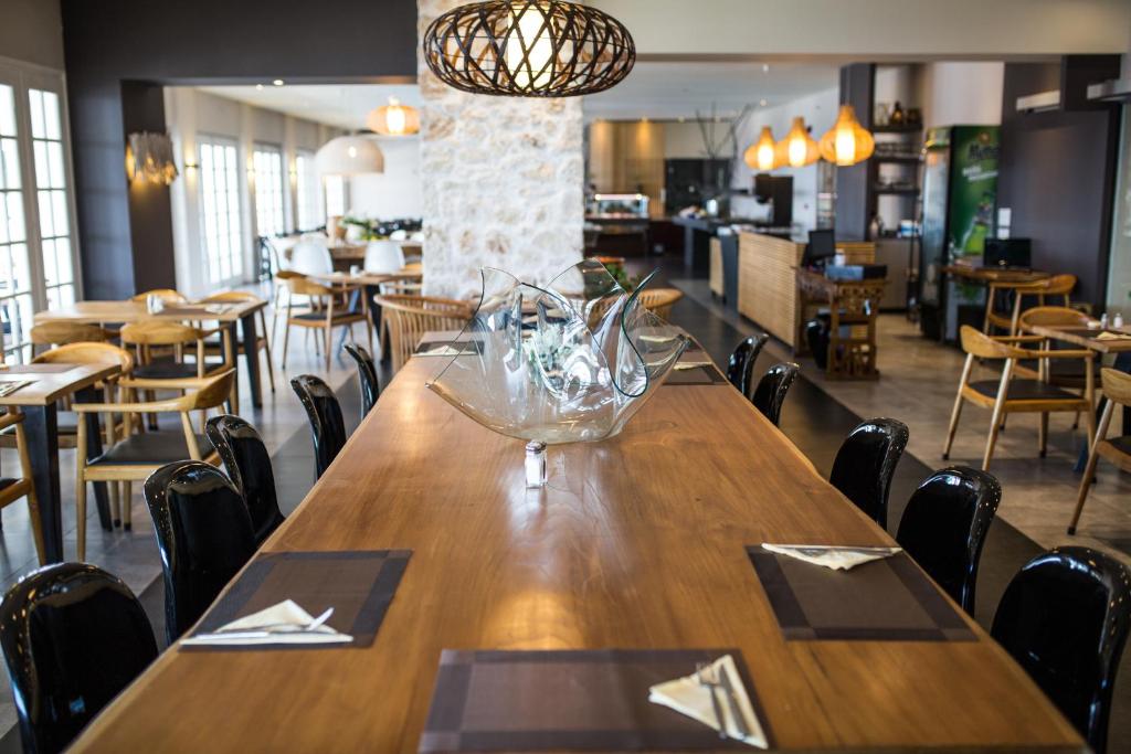 Mūsdienīgs restorāns ar plašu koka galdu, elegantu dekoru un stilīgām gaismām, kas rada siltu un aicinošu atmosfēru. Redzamas arī ēdamvietas un restorāna interjers.