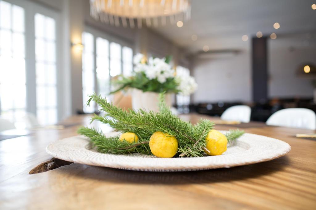 Dekoratīva plate ar zaļām priedes zariem un dzeltenām augļiem, novietota uz koka galda. Fons satur gaiši apgaismotu telpu ar logiem un ziediem.