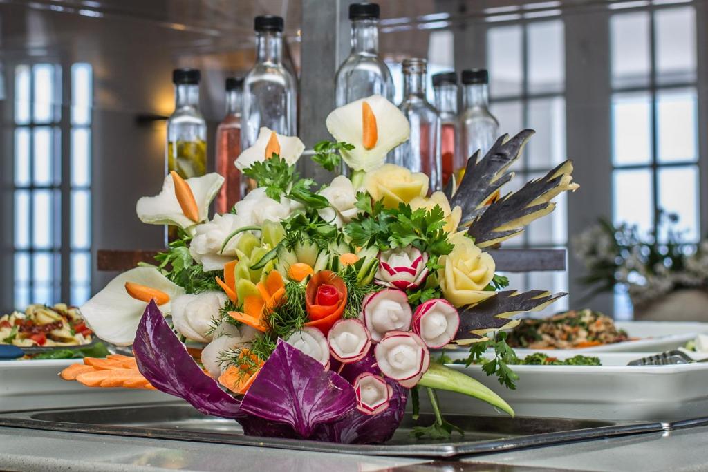 Svaigu dārzeņu kompozīcija ar dažādiem dārzeņiem un garšaugiem, radot krāsainu un pievilcīgu izskatu. Fons ar glāzēm un ēdieniem papildina atmosfēru.