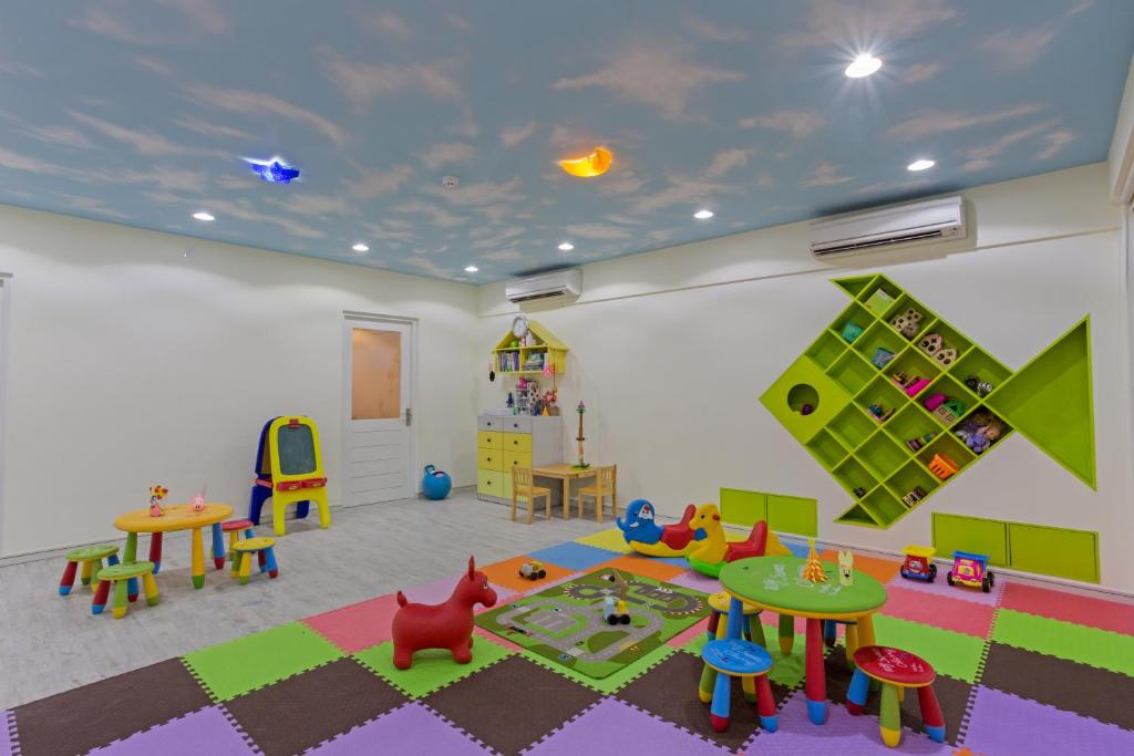 Krāsaina bērnu rotaļu istaba ar dažādām spēlēm, galdiem un krēsliem, kā arī dekoratīvu zilo debesu griestu dizainu. Istabā ir arī uzglabāšanas plaukti un rotaļlietas.