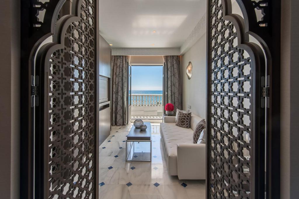 Eleganta viesnīcas numura skats ar jūras panorāmu, atverot durvis uz modernu interjeru ar gaisīgu viesistabu un komfortablu dīvānu.