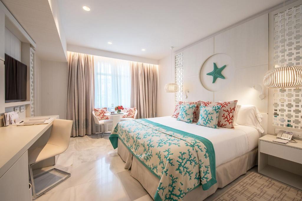 Mūsdienīga viesnīcas numura interjers ar baltām sienām un gaisīgiem aizkariem, grezns divguļamais gultas pārklājs ar jūras tematiku un ērtu sēdvietu stūrī.