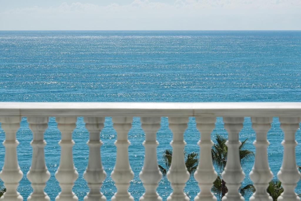 Skaists skats uz jūru no balkona, ar baltu balustrādi priekšplānā un palmu kokiem tuvumā.