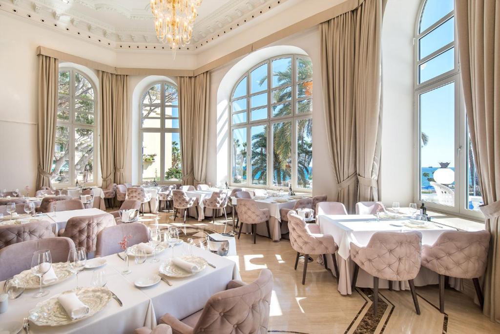 Elegants restorāns ar panorāmas logiem, kas piedāvā skatu uz jūru. Interjerā ir gaišas krāsas un grezni galdi ar izsmalcinātiem galda piederumiem.