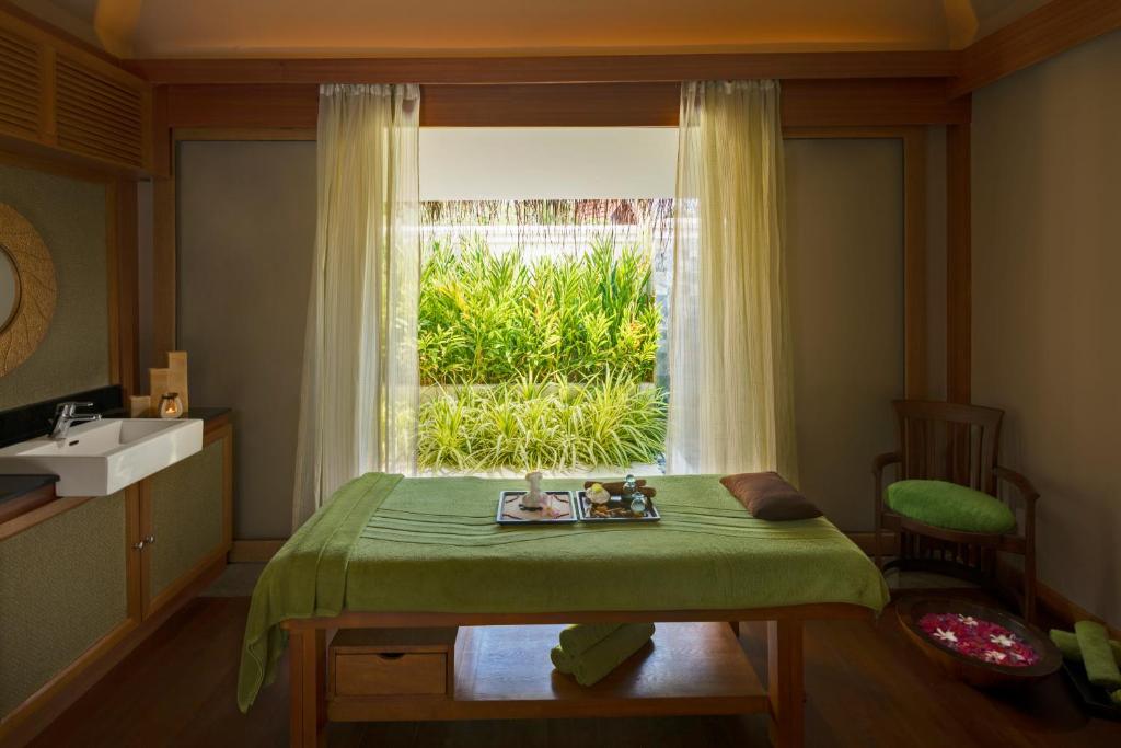 Spa telpa ar masāžas galdu, zaļu palagu un skatu uz dārzu caur lieliem logiem. Telpā ir dizaina elementi, kas rada relaksējošu atmosfēru.