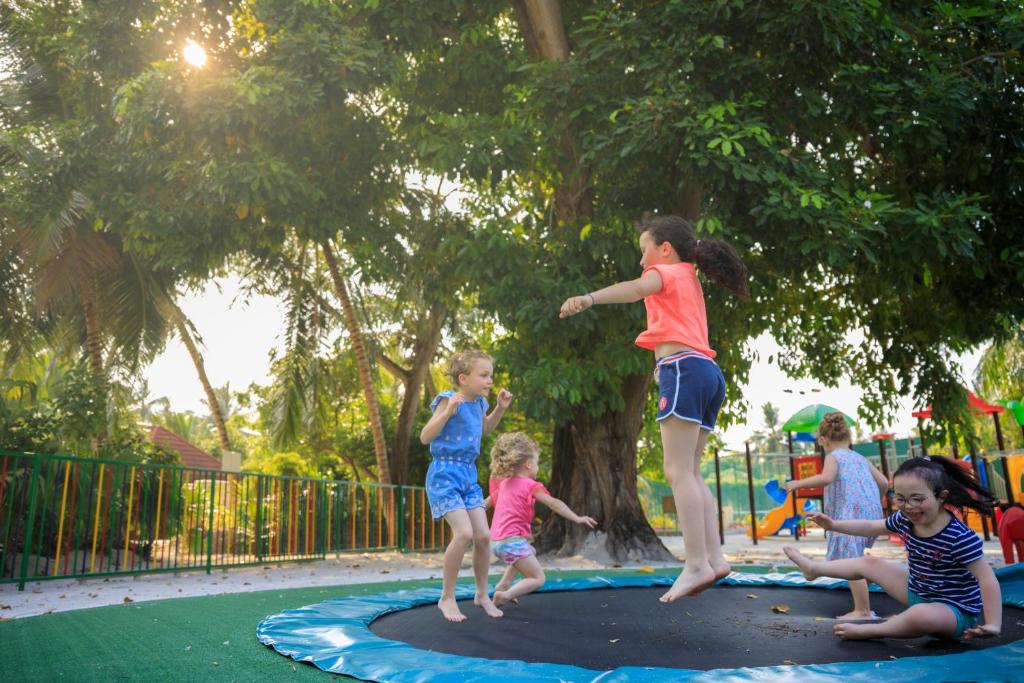 Bērni jautri lēkā uz batuta saulainā parkā, apkārt zied zaļumi un spēļu laukums.