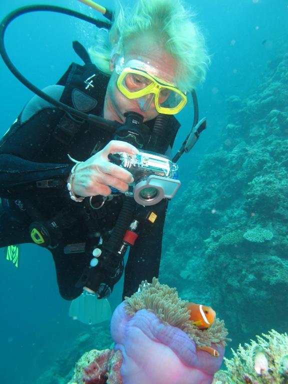 Aptverta nirēja, kas fotografē zemūdens pasauli, tuvplānā redzama anemone ar clownfish. Ūdens ir skaidrs, un apkārt ir korallriffi.