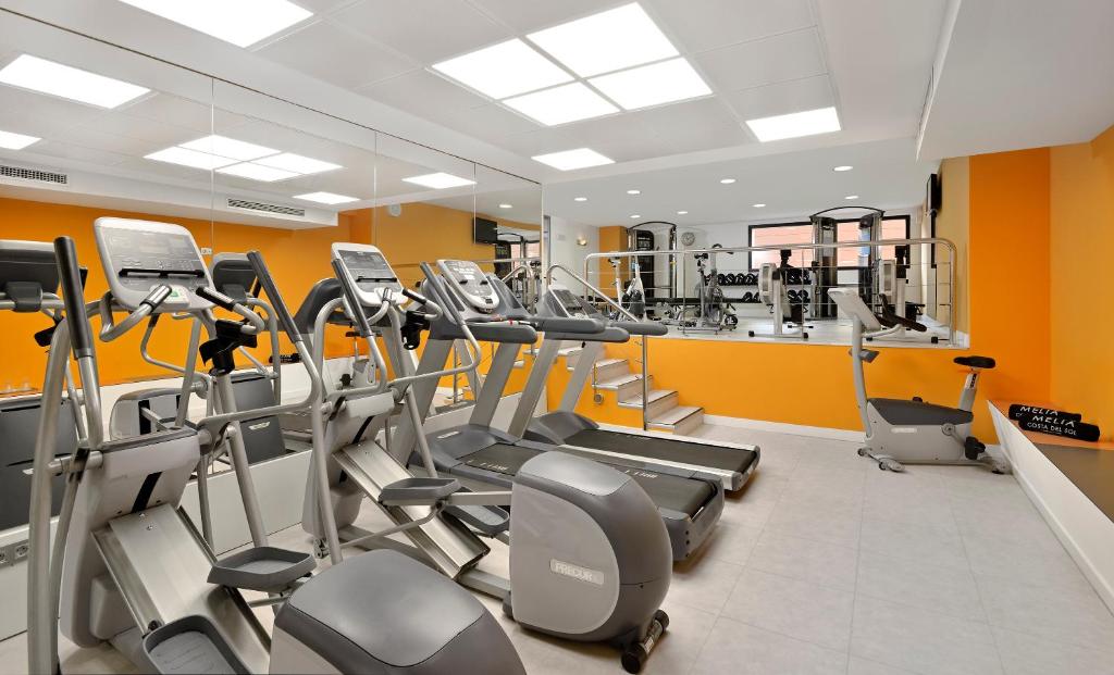Mūsdienīgs trenažieru zāles interjers ar dažādiem fitness aprīkojumiem, tostarp skrejceliņiem un eliptiskajiem trenažieriem, ar gaišu apgaismojumu un dzīvīgu oranžu sienu.