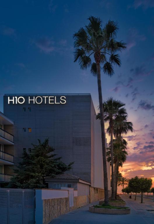 H10 Hotels logo redzams pie viesnīcas ēkas, apkārt palmas un krāsaina saulrieta ainava.