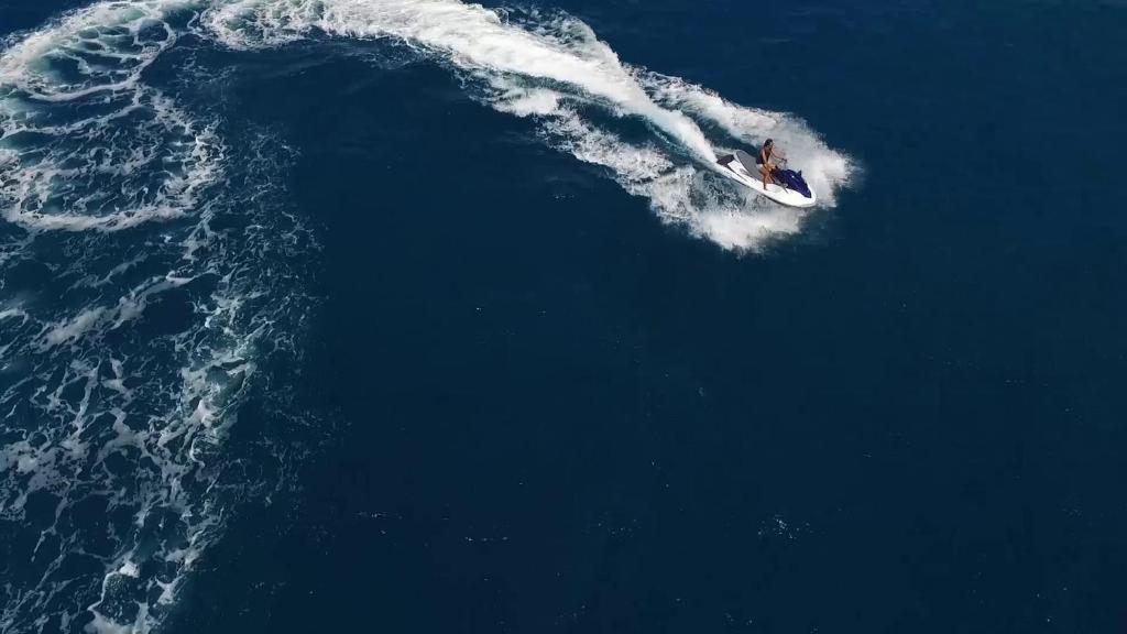 Personāžs izpilda akrobātiskus trikus uz ūdens motocikla, radot viļņus zilajā jūrā. Daba un ūdens sports apvienojas aizraujošā mirklī.