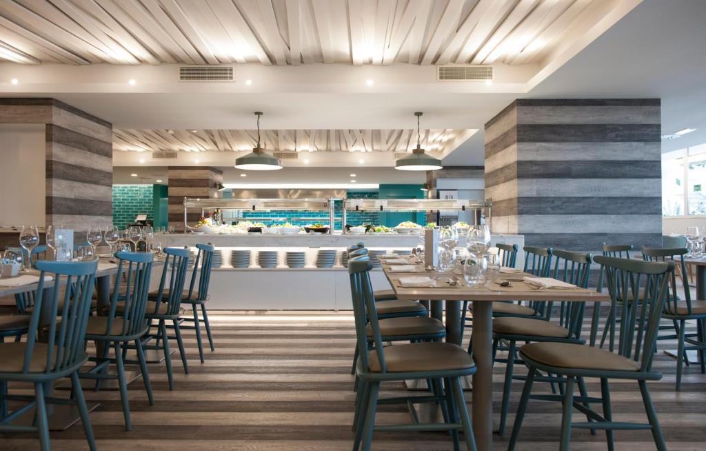 Mūsdienīgs restorāna interjers ar koka grīdu un stilīgiem, gaiši ziliem krēsliem. Vērienīga bufete ir redzama fona, nodrošinot viesiem ērtu piekļuvi ēdieniem.